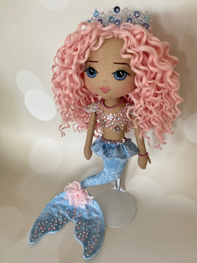 Mermaid Doll with blue tail, pink and silver diamante detail and mermaid bra. Curly hair made from hand rolled pink mohair fibre and a mermaid crown in silver with blue sequin detail. Doll features a hand embroidered face featuring blue eyes, pink lips and three dimensional eye lashes
