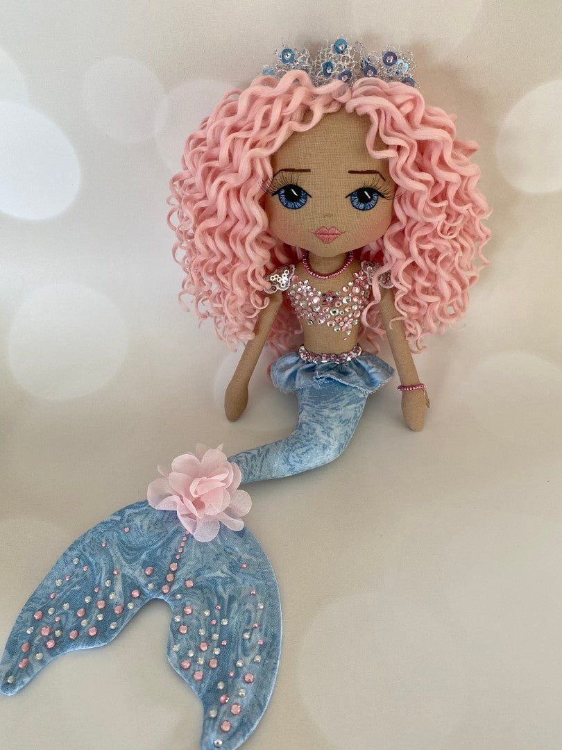Mermaid Doll with blue tail, pink and silver diamante detail and mermaid bra. Curly hair made from hand rolled pink mohair fibre and a mermaid crown in silver with blue sequin detail. Doll features a hand embroidered face featuring blue eyes, pink lips and three dimensional eye lashes.