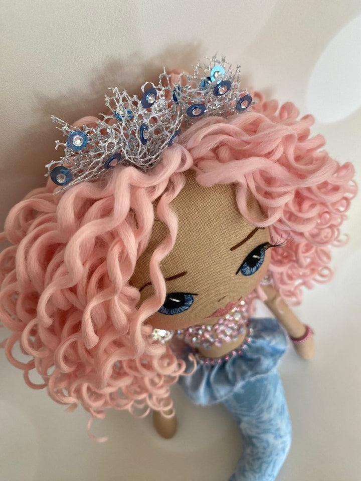 Mermaid Doll with blue tail, pink and silver diamante detail and mermaid bra. Curly hair made from hand rolled pink mohair fibre and a mermaid crown in silver with blue sequin detail. Doll features a hand embroidered face featuring blue eyes, pink lips and three dimensional eye lashes.