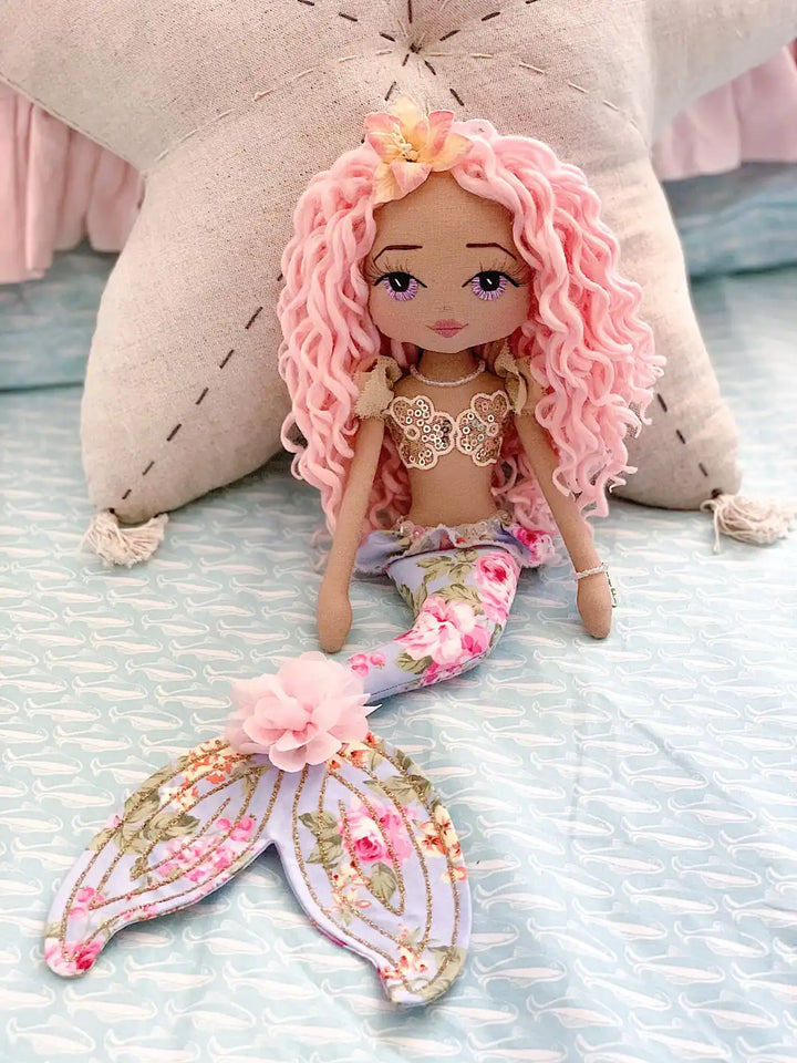 mermaid doll with long pink curly hair, hand embroidered lavender eyes, gold sequin mermaid top, blue floral mermaid tail with gold glitter detail.
