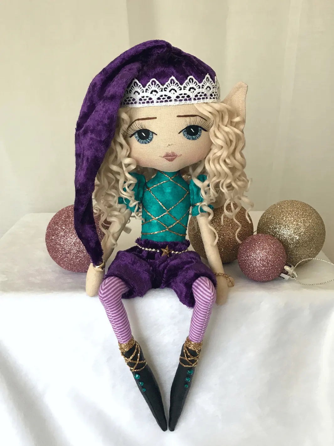 handmade christmas elf doll sitting on a shelf wearing purple velvet hat & pants, purple striped tights, teal & gold elf top, long blond curly hair, hand embroidered blue eyes