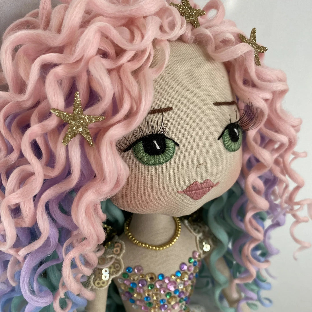 Close-up of the Kalani Auralis mermaid doll’s face, featuring embroidered green eyes, soft pink lips and pastel rainbow curls.