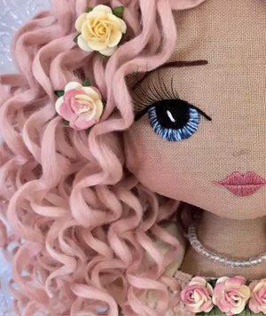close up image of an Upper Dhali handmade doll with hand embroidered face featuring blue eyes, 3d eyelashes, curly pink hair adorned with light yellow and pink roses