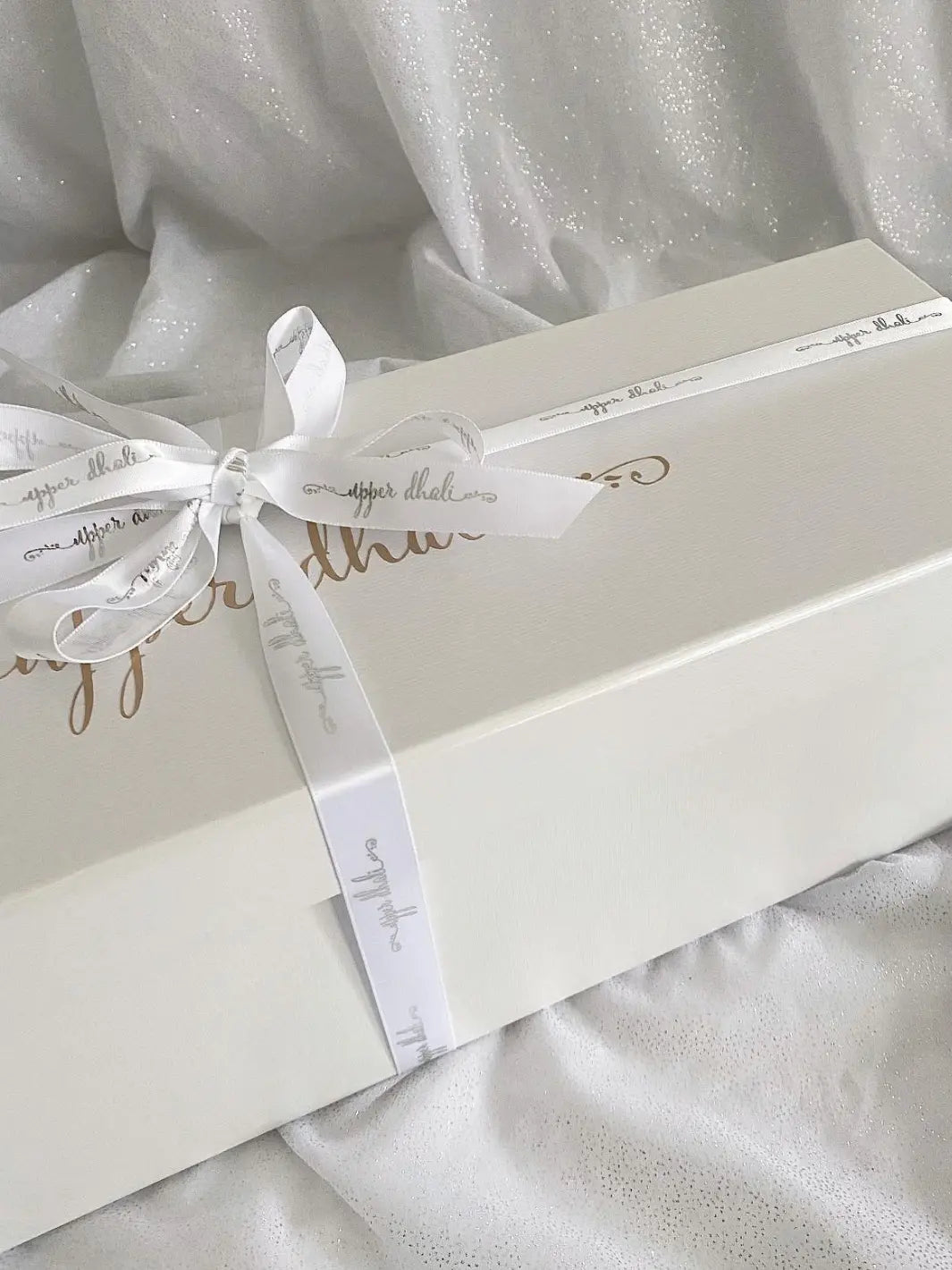 close up image of white gifting box with gold logo & white satin ribbon for Upper Dhali handmade dolls