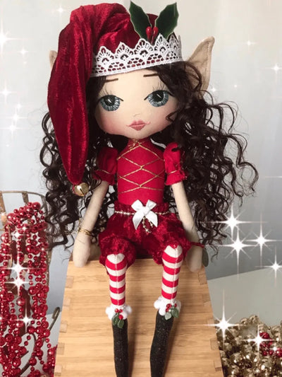 handmade christmas elf sitting on a wooden shelf wearing a red and white santa inspired outfit. Red velvet pants & elf jingle bell hat, long brown curly hair, hand embroidered blue eyes and holly embellishments