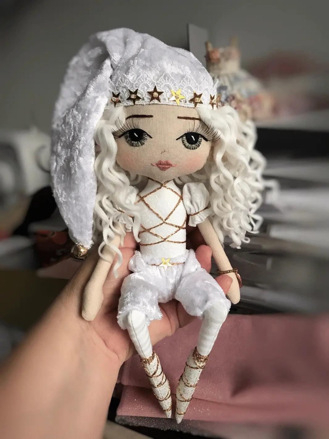 handmade christmas elf with bended legs to sit on a shelf, wearing white and gold outfit with hand embroidered green eyes; being held in a woman's hand