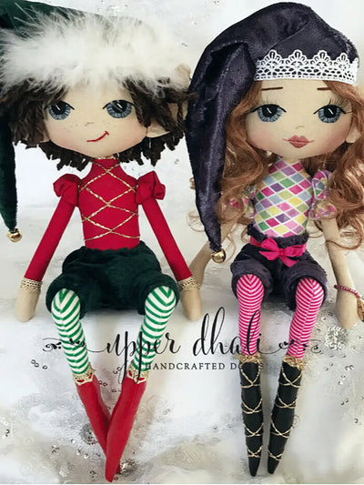 two handmade christmas elf dolls, one boy and one girl, sitting side by side on a shelf. The boy is wearing a green velvet santa hat, red top with gold embellishments, green velvet pants, green and white striped tights and red shoes. The girl is wearing a purple velvet santa hat, with a multicoloured top, purple velvet pants and pink striped tights. Both have hand embroidered blue eyes.