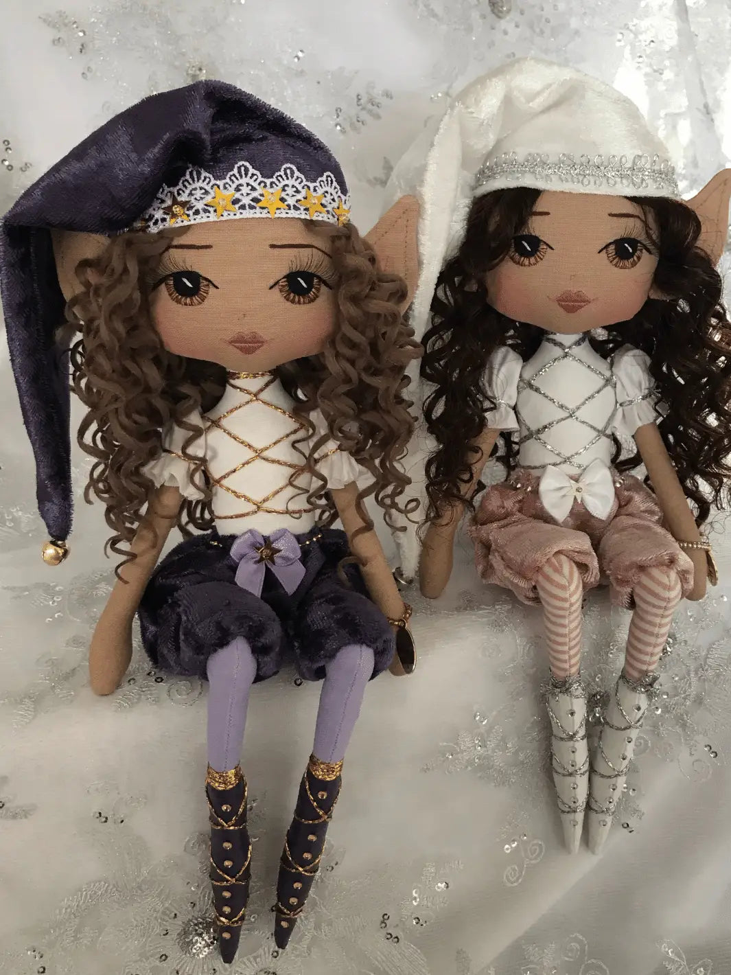 two cheeky christmas elf handmade dolls sitting on a shelf. One is purple tones with brown eyes and brown curly hair, the other is white with pink pants, long dark curly hair and brown eyes