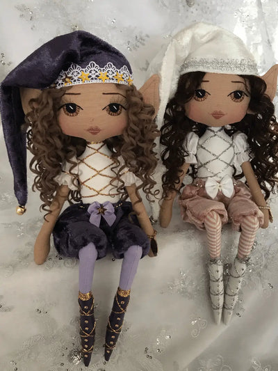 two cheeky christmas elf handmade dolls sitting on a shelf. One is purple tones with brown eyes and brown curly hair, the other is white with pink pants, long dark curly hair and brown eyes