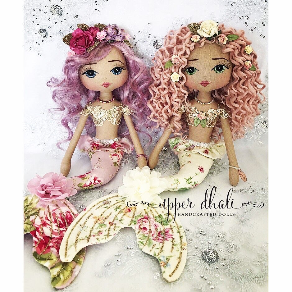 two mermaid dolls sitting side by side. one has purple hair and a purple floral tail, the other has a cream floral tail and pink curly hair