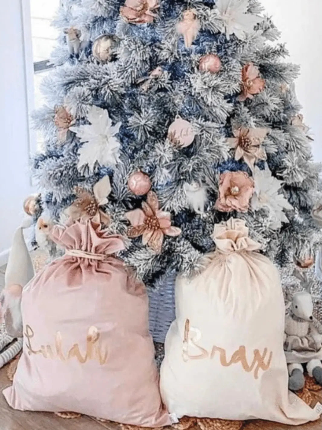 two velvet personalised santa sacks, one pink, one ivory, sitting in front of a beautifully decorated christmas tree adorned with white and pink decorations