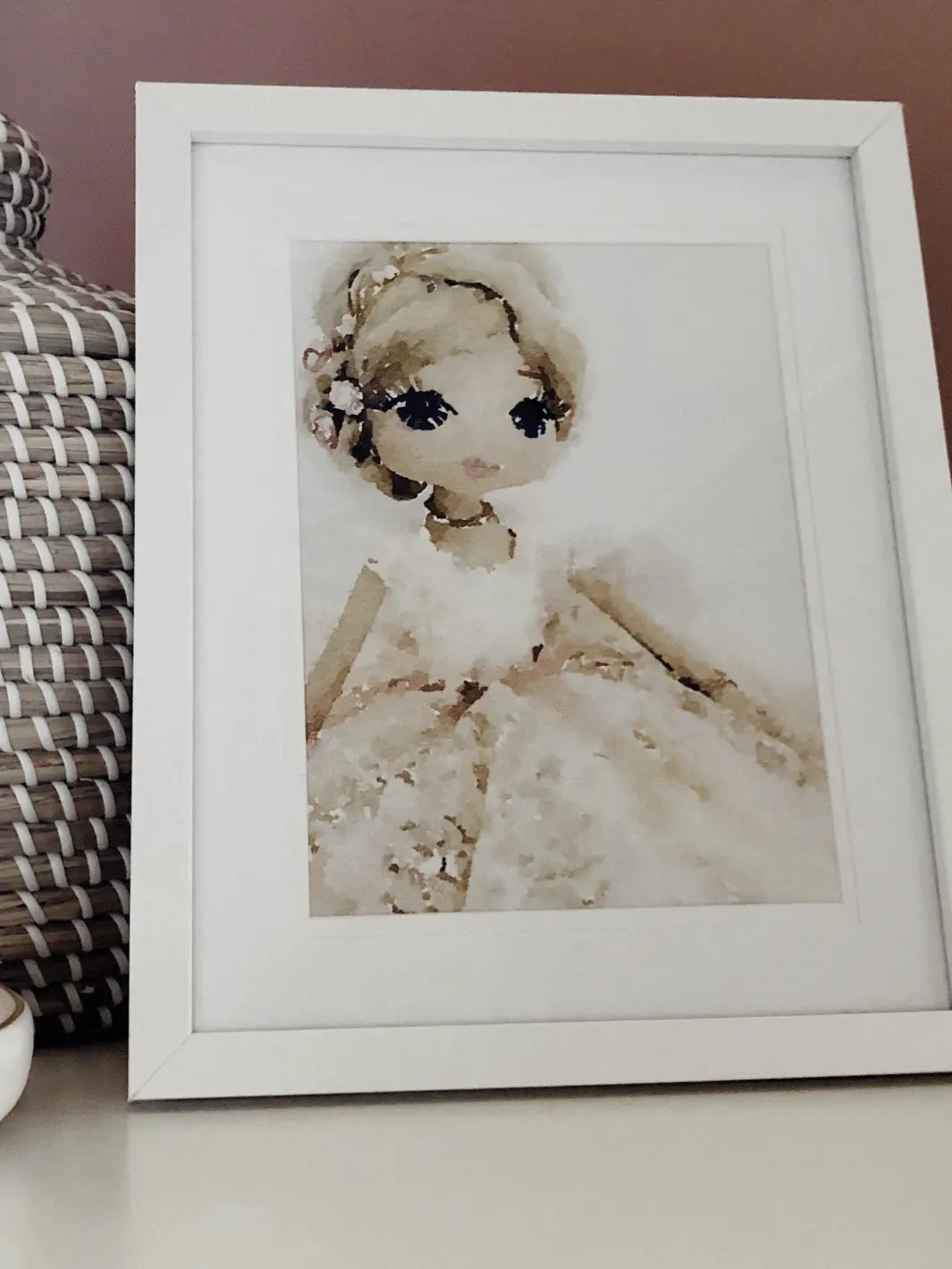 watercolour print of handmade doll in neutral and pink tones in a frame displayed on a bench