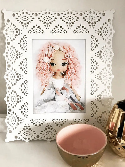 watercolour art print of a handmade doll with pink curly hair and a white and blue dress, displayed in a white frame on a bench beside a gold and pink bowl and a gold vase